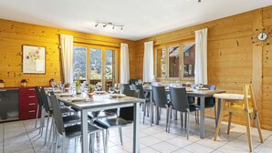 Un espace repas avec une grande table dressée pour six personnes, entourée de chaises, et offrant une vue sur une forêt grâce à de grandes fenêtres.
