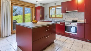 Une cuisine moderne dotée d’un îlot central, de meubles de rangement rouges et offrant une vue sur un paysage verdoyant grâce à de grandes fenêtres.