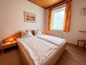 Een tweepersoonsbed met witte lakens, een nachtkastje met lamp, een schilderij aan de muur, en een raam met gordijnen.