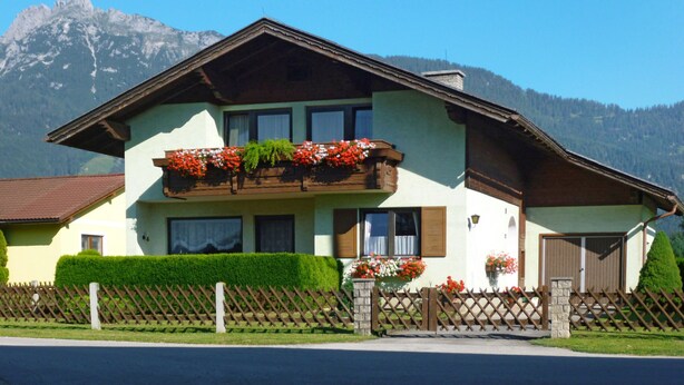 Une maison à deux étages, dotée d’un toit en pente, d’un balcon en bois orné de jardinières et d’une clôture en pierre.