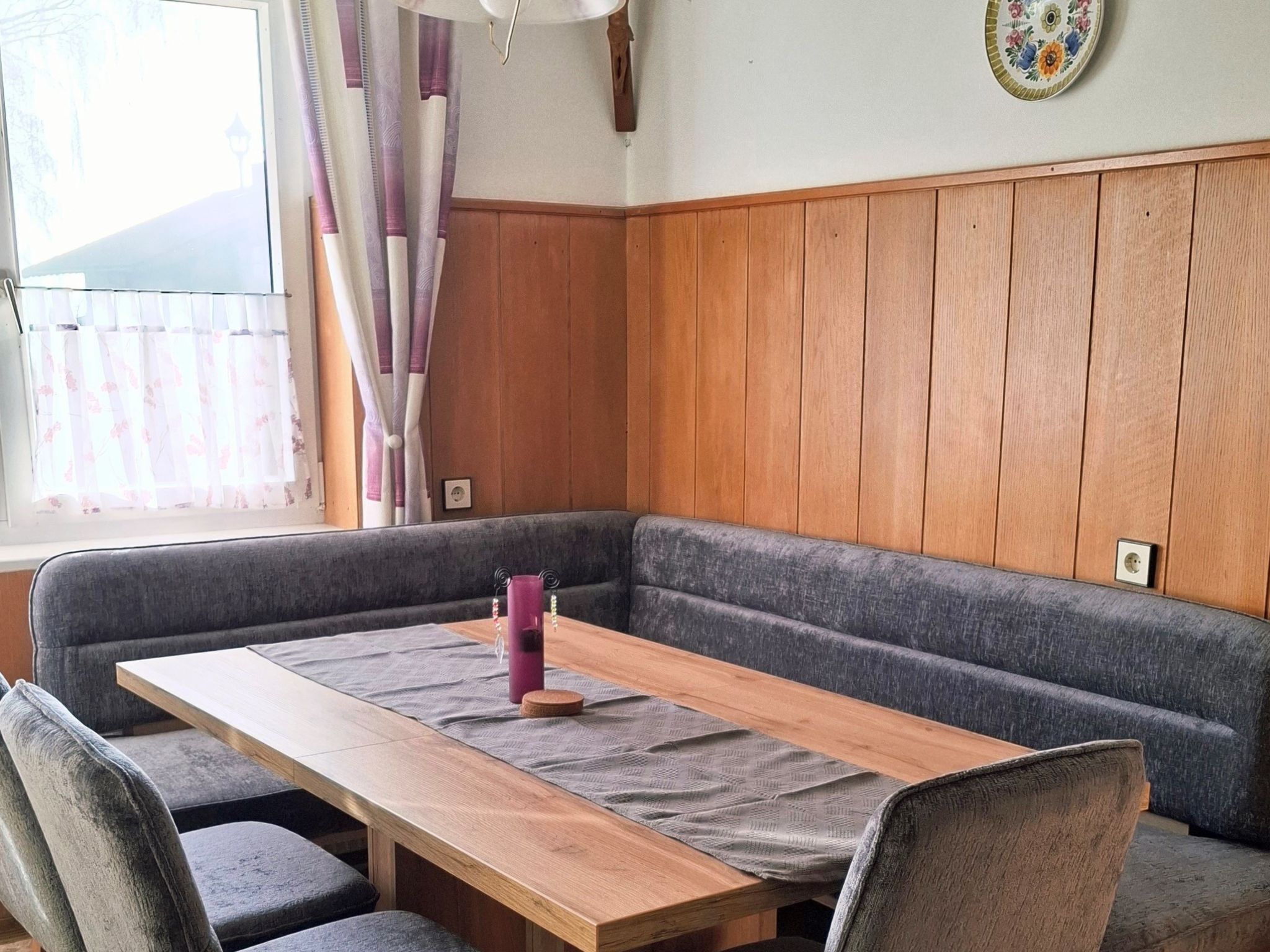 A cozy room with a wooden table, grey upholstered seating, and a window with curtains.