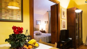 A room with a dining table set with fruit, a potted plant, and a mirror reflecting a bed and a lamp.