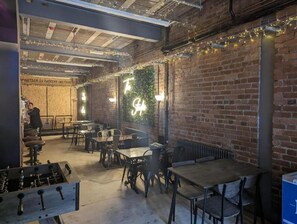 Restaurante com paredes de tijolo, luminárias pendentes e uma placa de neon. Mesas e cadeiras dispostas para refeições.