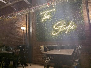 A restaurant interior with a neon sign reading "The Studio" on a brick wall, metal chairs, and tables. - The Studio (Blackpool)