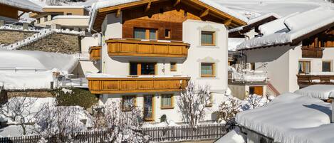 A multi-story chalet with wooden balconies, surrounded by snow-covered trees and a clear blue sky.