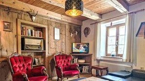 Um quarto acolhedor com paredes de madeira, uma estante de livros, dois poltronas de veludo vermelho, um sofá de couro preto e uma televisão.