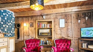 Um quarto aconchegante com paredes de madeira, duas poltronas vermelhas macias, uma estante de livros, um rádio antigo e uma televisão.