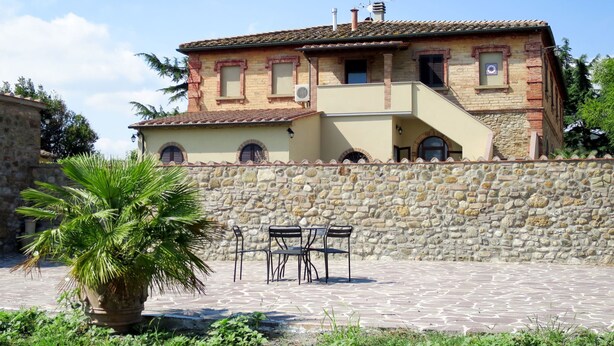 Ein zweigeschossiges Haus mit einer Steinmauer, einem Ziegeldach und einer kleinen Terrasse mit Stühlen und einem Tisch.