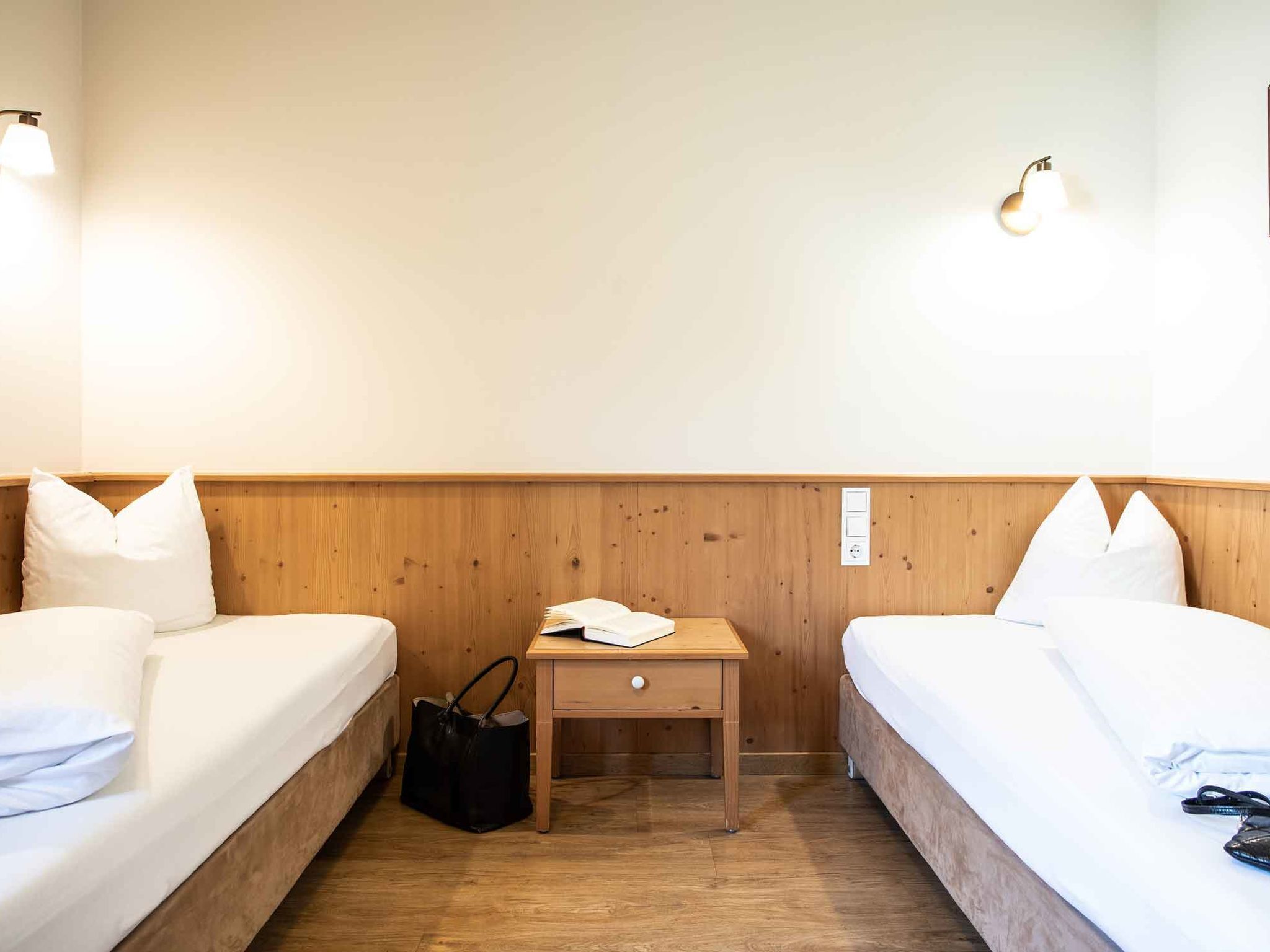 A hotel room with two single beds, a wooden nightstand with a lamp, and a small table with a book and a bag.