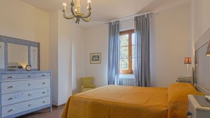 A bedroom with a bed, a dresser, a chair, a window with curtains, and a chandelier. - Casa alle Vacche by Interhome (San Gimignano)