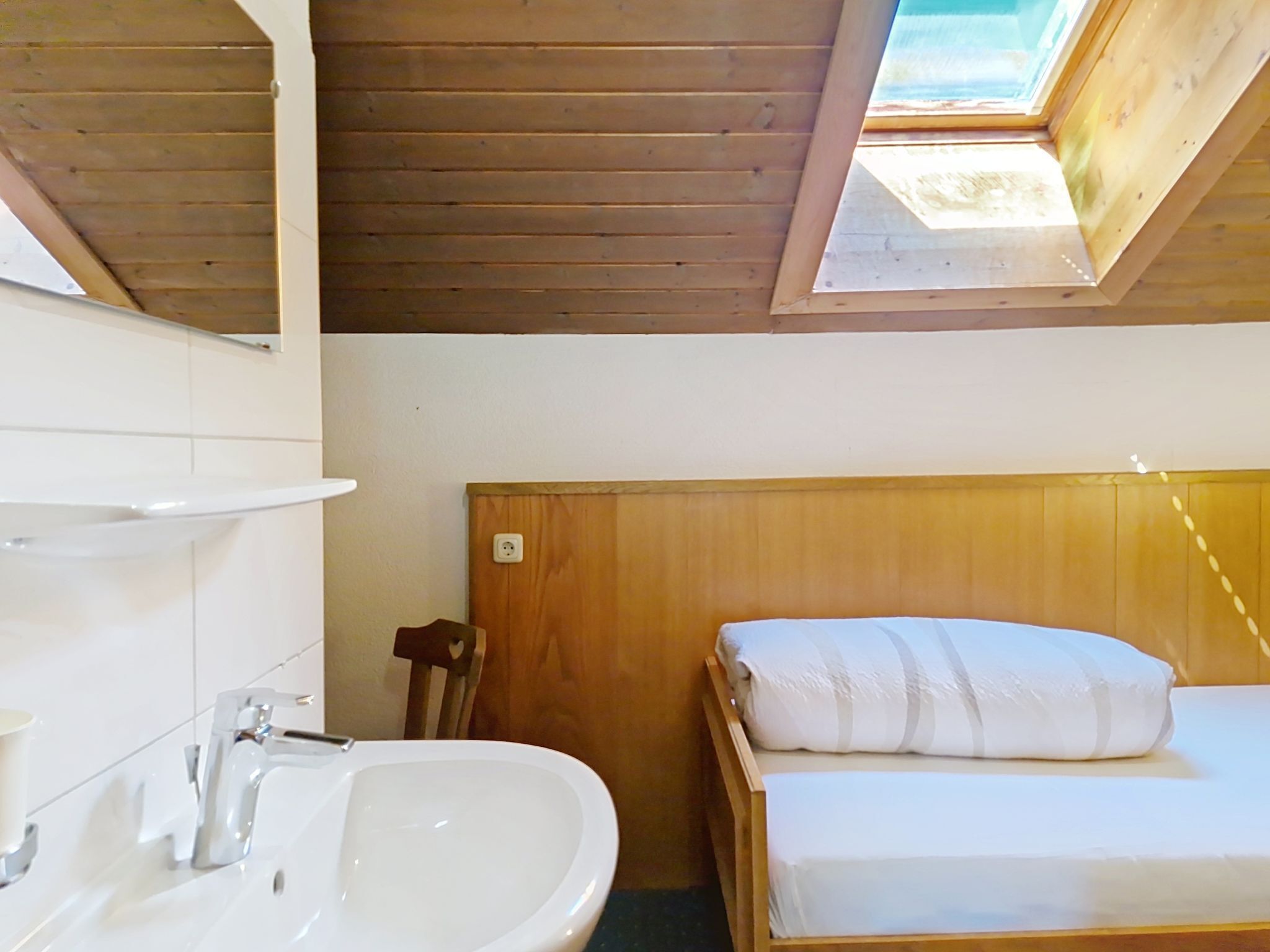 A small, cozy room with a skylight, a wooden headboard, a white bed, and a sink with a mirror.