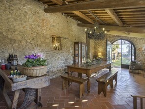 Un intérieur rustique avec des murs en pierre, des poutres en bois, une grande table avec des bancs et une vue sur l’extérieur, visible à travers une porte en arc de cercle.