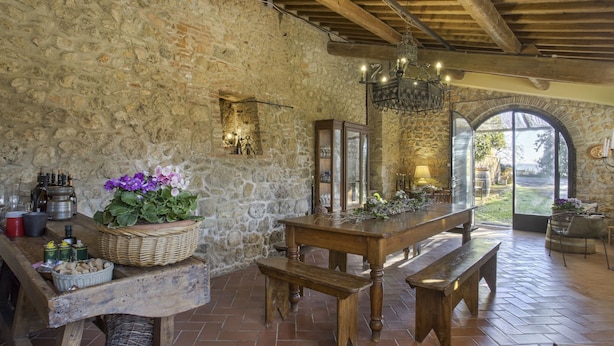 Un interior rústico con paredes de piedra, vigas de madera, una mesa grande con bancos y una vista a través de un arco.