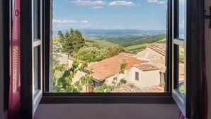 Vista desde una ventana sobre un paisaje rural con colinas onduladas, cielo despejado y edificios tradicionales de piedra con tejados de tejas rojas.