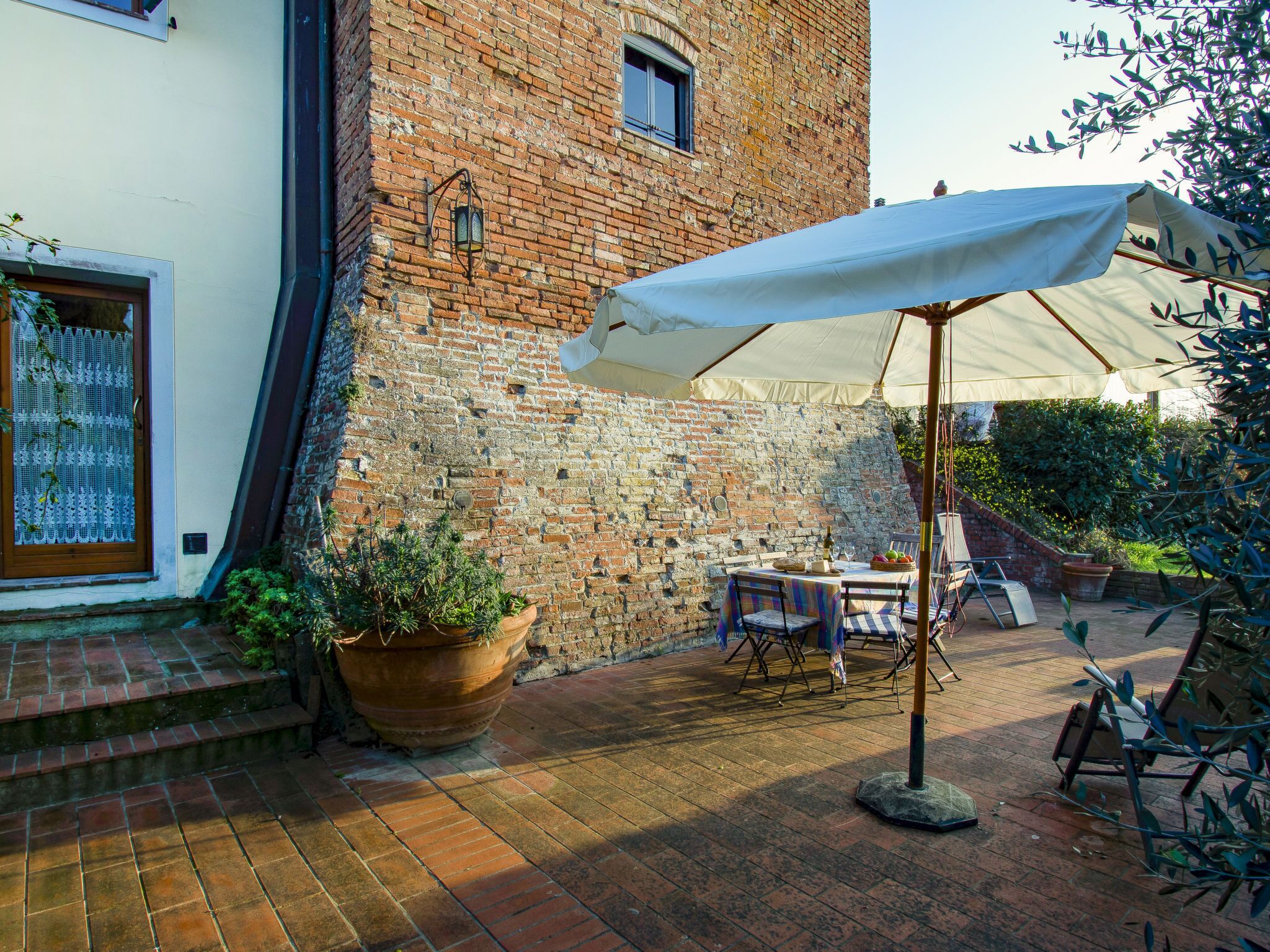 Un patio al aire libre con pared de ladrillo, una mesa con sillas, un paraguas grande y plantas en macetas.