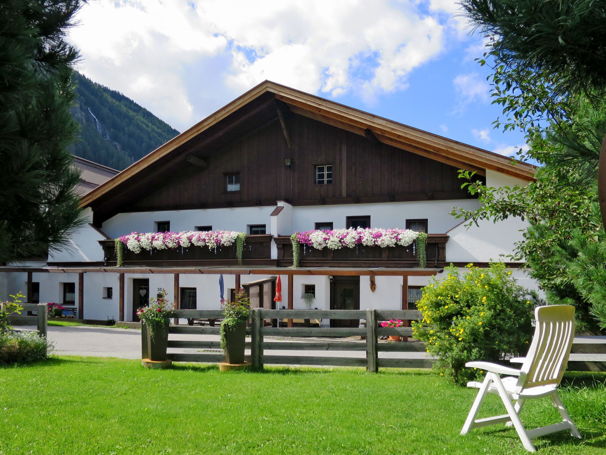 A traditional chalet with a wooden facade, balconies with flower boxes, and a grassy foreground with a bench and chair.
