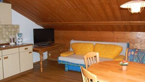 Tunnelmallinen ullakkohuone, jossa on puinen katto, kuviollisilla tyynyillä varustettu sohva, televisio, kahvinkeitin ja ruokapöytä tuoleineen.