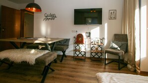 Ein gemütliches Zimmer mit einem Holztisch, einer Couch und einem Stuhl. An der Wand hängt ein Fernseher und ein Schild mit der Aufschrift 'Guten Appetit'.