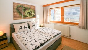 Ein Schlafzimmer mit Bett, Nachttisch, Wandbild, Fenster mit Vorhängen und Heizkörper.