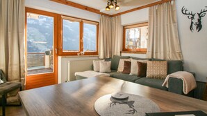 Ein gemütliches Wohnzimmer mit einem Holztisch, einer Couch mit gemusterten Kissen und Blick auf eine Berglandschaft durch große Fenster.