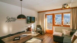 Ein modernes Wohnzimmer mit einem Holzesstisch, einer Couch und Blick auf eine Berglandschaft durch große Fenster.