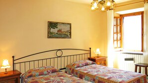 A bedroom with two beds, a nightstand with a lamp, a dresser, a painting on the wall, and a window with curtains. - Castagni II (OCT101) by Interhome (Orciatico)