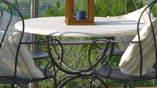A round table with a lantern on it, surrounded by metal chairs with cushions, set on a terrace with a view of green fields and hills. - Castagni I (OCT100) by Interhome (Orciatico)
