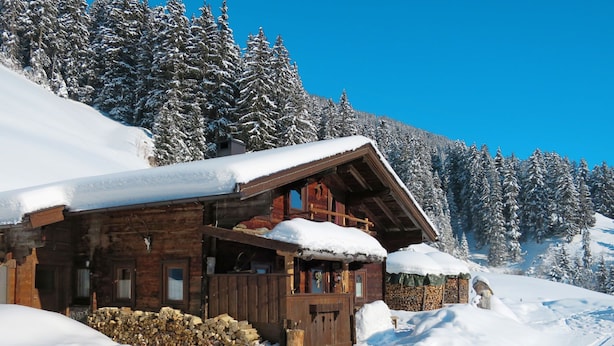 Eine Holzhütte mit geneigtem Dach, umgeben von schneebedeckten Bäumen und blauem Himmel.