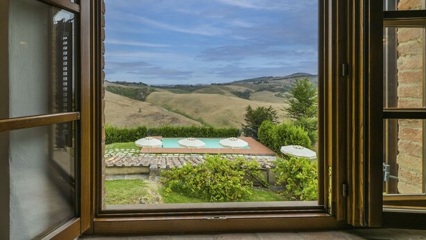 On aperçoit, à travers une fenêtre ouverte, un espace piscine avec une table et des parasols, le tout entouré de verdure et avec des collines en arrière-plan.