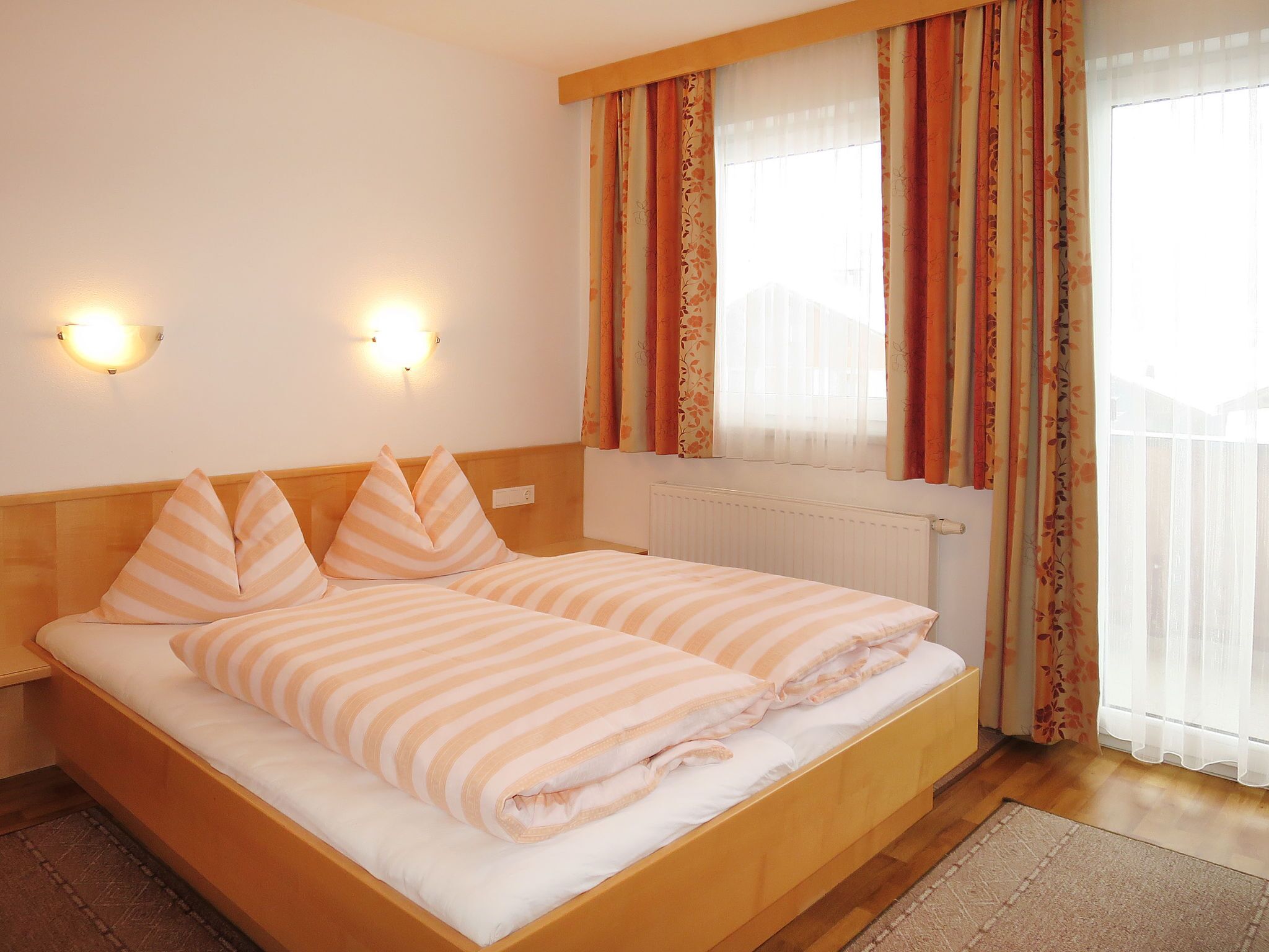 Cama doble con ropa de cama a rayas en una habitación con cabecera de madera, dos lámparas de pared y un ventanal con cortinas.