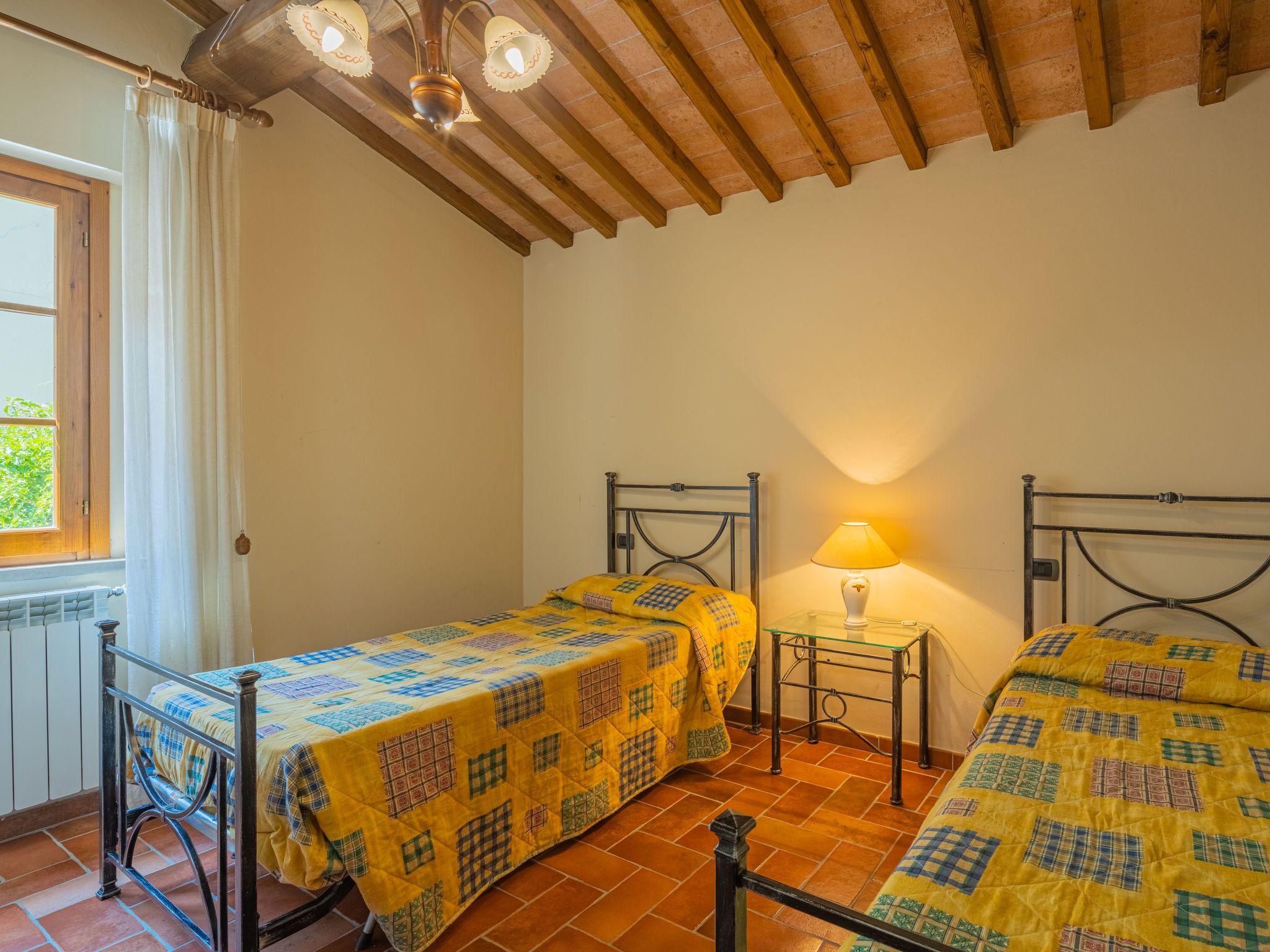 A room with two beds, each with a metal frame and yellow bedding. There is a small bedside table with a lamp between the beds. The room has a tiled floor and a wooden ceiling with exposed beams.