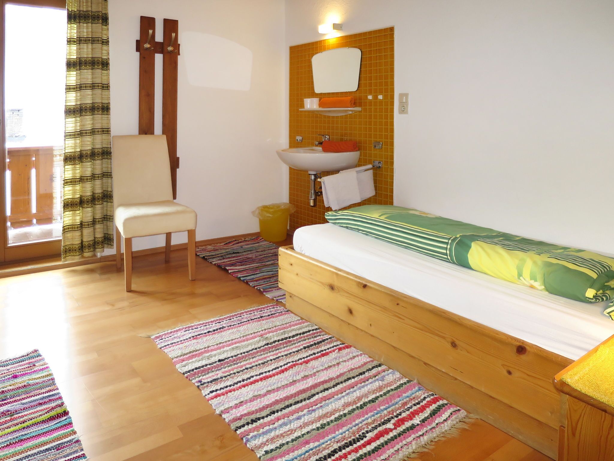 Quarto com cama de madeira, uma cadeira, pia pequena e janela com cortinas.
