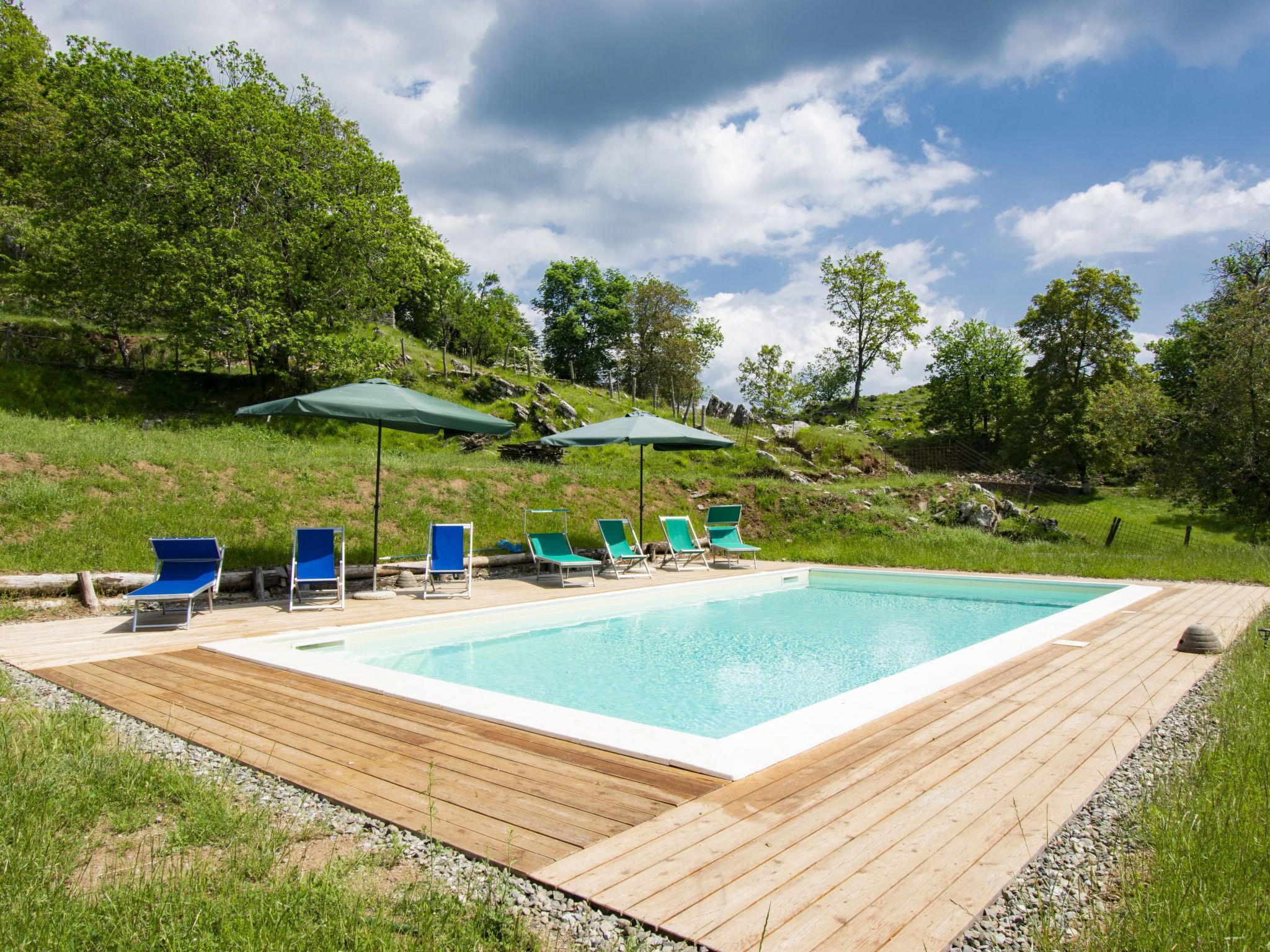 Una piscina rettangolare con un pontile in legno, circondata da sedie a sdraio verdi e ombrelloni.
