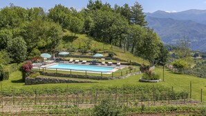 A pool area with lounge chairs and umbrellas, surrounded by a stone wall and lush greenery, with mountains in the background. - Le Bore (CNG122) by Interhome (Castelnuovo di Garfagnana)