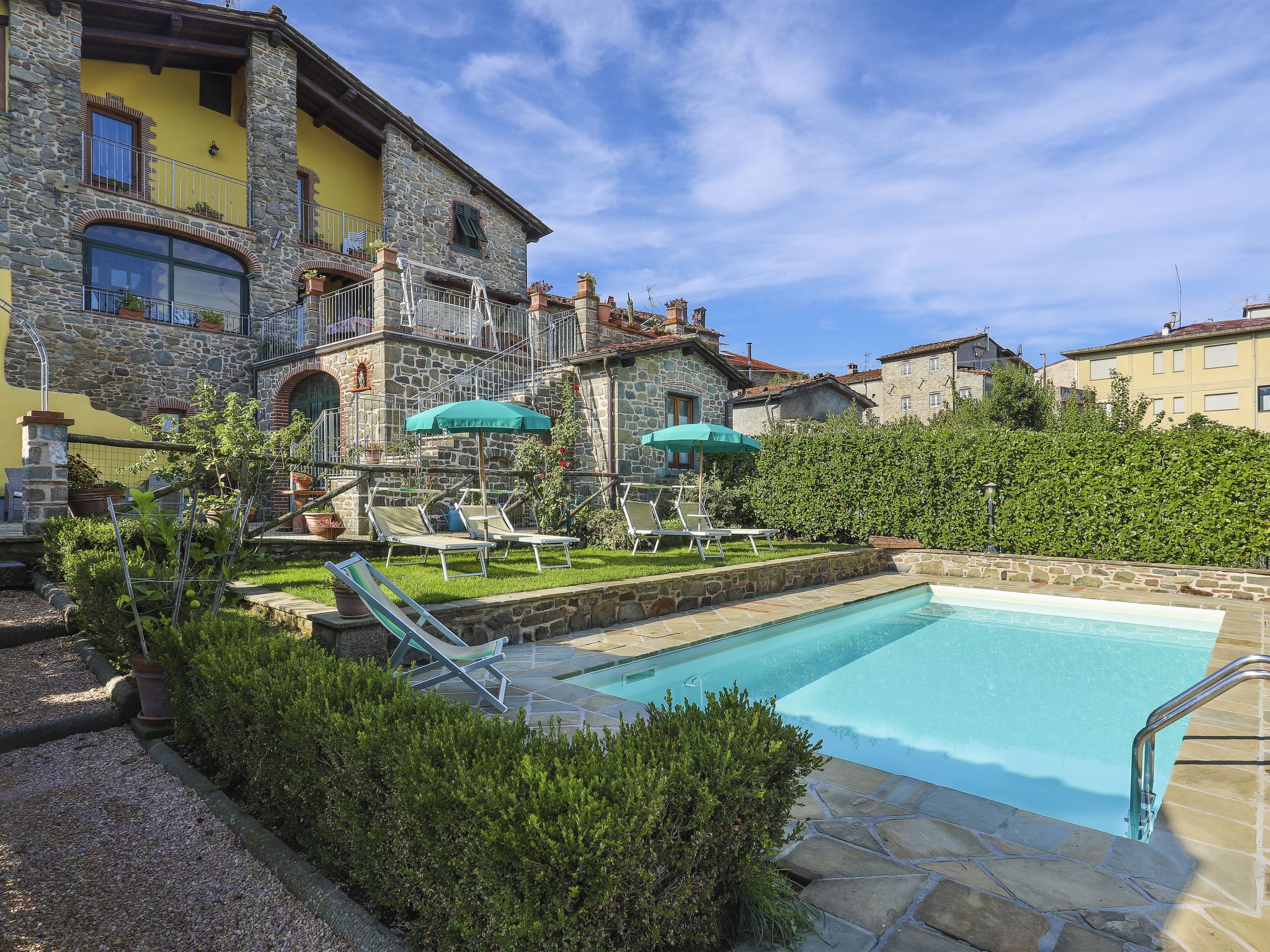 A pool area with stone walls, a stone building with balconies, and a hedge.