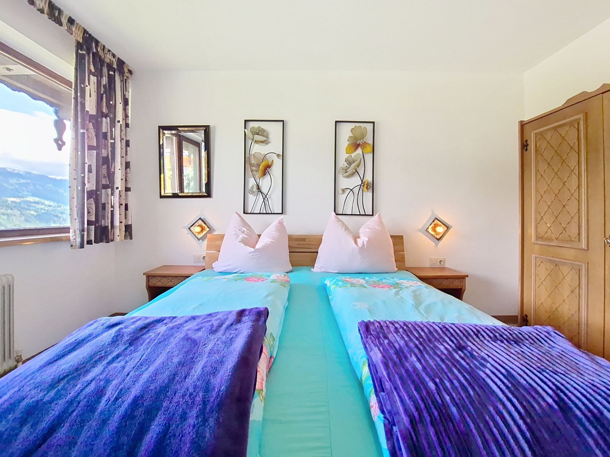 A double bed with floral bedding, two bedside tables, a window with curtains, and wall art.