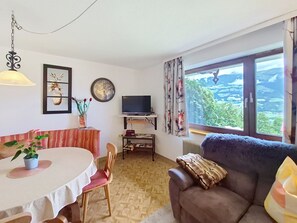 A living room with a sofa, a dining table with chairs, a TV, a clock, and a window with a view. - Höllwarth by Interhome (Fügen)