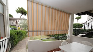 Un balcó amb una taula i cadires blanques, una cortina estriada i vista a un jardí i edificis veïnals.