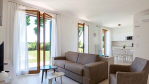 A modern living room with a sofa, a coffee table, and a chair. There is a kitchen area with a microwave and a dining table. Large windows provide natural light and a view of greenery outside. - Ca' Del Pioppo by Interhome (Cavallino)