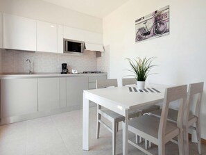 Une cuisine moderne avec des meubles blancs, un four à micro-ondes et une machine à café. On y trouve une table à manger avec des chaises et une plante en pot.