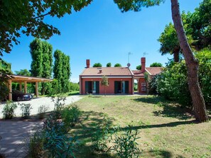 Une maison à deux étages, avec un toit recouvert de tuiles rouges, une terrasse couverte et un jardin agrémenté de diverses plantes et arbres.