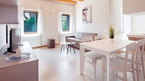 Eine moderne Küche mit weißen Schränken, ein Essbereich mit weißem Tisch und Stühlen sowie eine Wohnbereich mit grauem Sofa und einer Pflanze.
