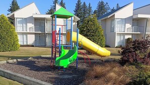 A playground with a slide and climbing structure, surrounded by a landscaped garden and residential buildings in the background.