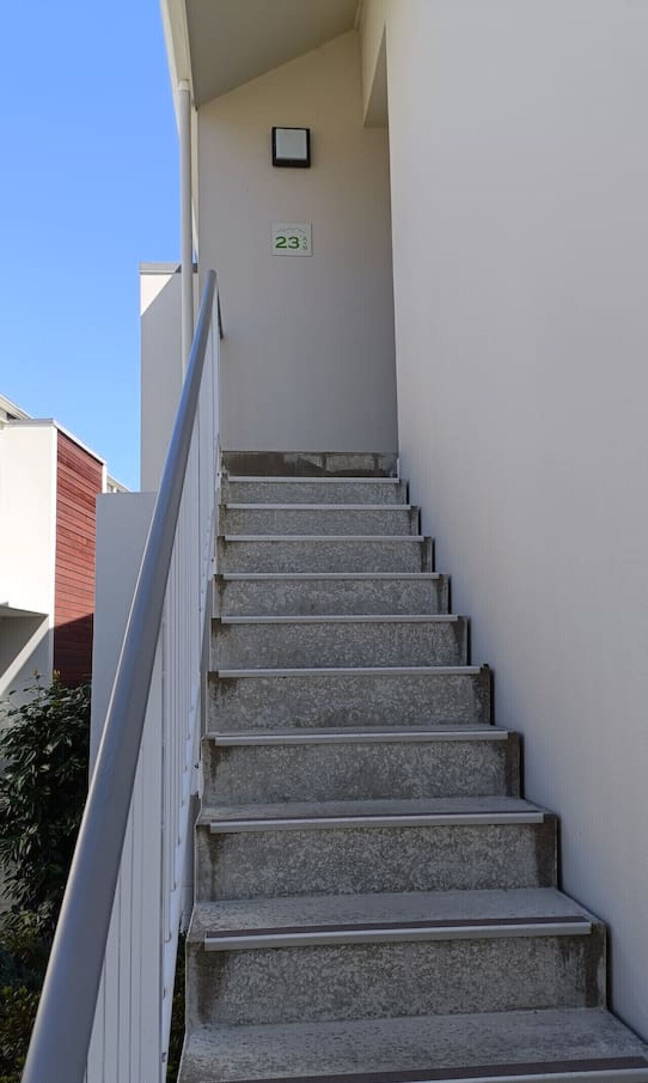 Uma escadaria com corrimão, uma porta com placa de número e céu claro.