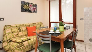 Un salon chaleureux avec un canapé, une table à manger en bois et des chaises. Il y a une fenêtre avec des rideaux, un tableau au mur et une plante en pot sur la table.