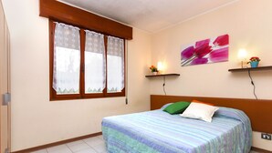 Une chambre à coucher avec un lit, une fenêtre avec des rideaux, une étagère fixée au mur ornée de fleurs et un tableau.