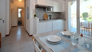 A modern kitchen with white cabinets, a microwave, and a sink. There is a dining area with a table set for two, and a view of the outdoors through a sliding glass door. - Elisabeth by Interhome (Lignano Pineta)
