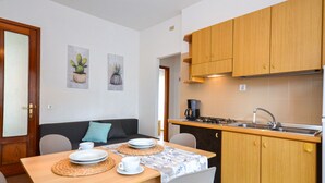 A compact kitchen with wooden cabinets, a sink, and a stove. There is a dining area with a table set for two, chairs, and a sofa. - Villa Rosanna by Interhome (Lignano Pineta)