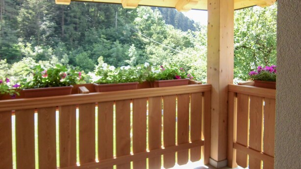 Een houten balkon met een bankje, omgeven door potplanten en uitzicht op een bebost gebied.