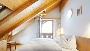 A bedroom with a wooden ceiling, a bed with white bedding, a chair, a bedside table with a book, and a window with curtains. - Ardoney by Interhome (Campitello)